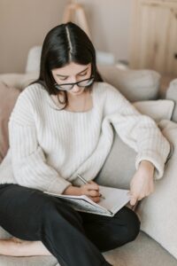 A young woman writes in a notebook while sitting comfortably on the sofa at home.