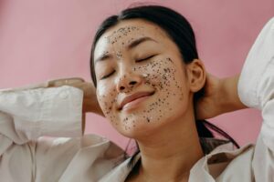 Asian woman with glitter face mask and eyes closed, experiencing a relaxing skincare routine against a pink background.