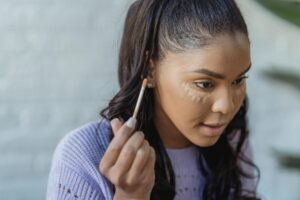 Crop African American woman with long dark hair applying concealer under eyes and looking away in light room