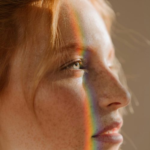 Elegant portrait of a redhead woman with rainbow light across her face, highlighting freckles and beauty.